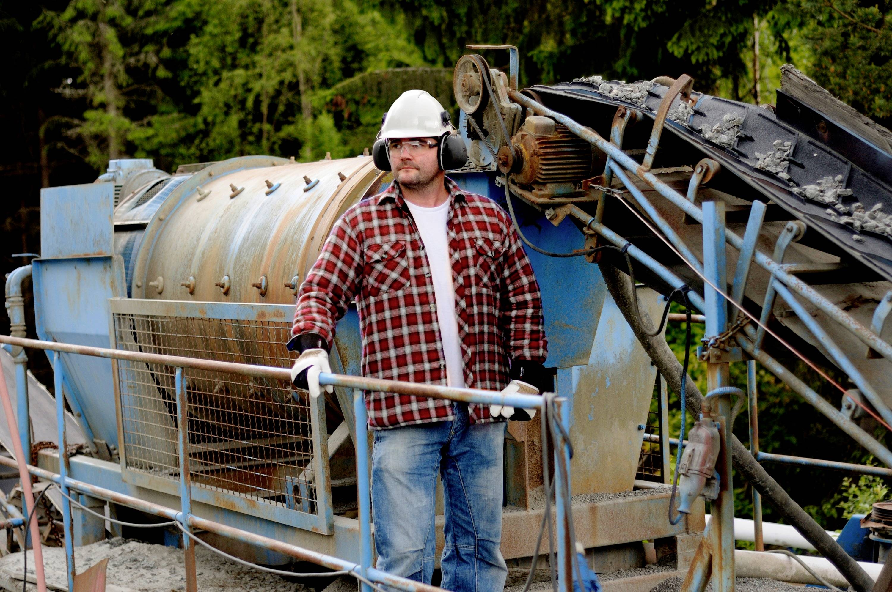 Ein Arbeiter mit Helm und Schutzkleidung steht neben einer großen Industriemaschine in einem Freiluft-Betriebsbereich.