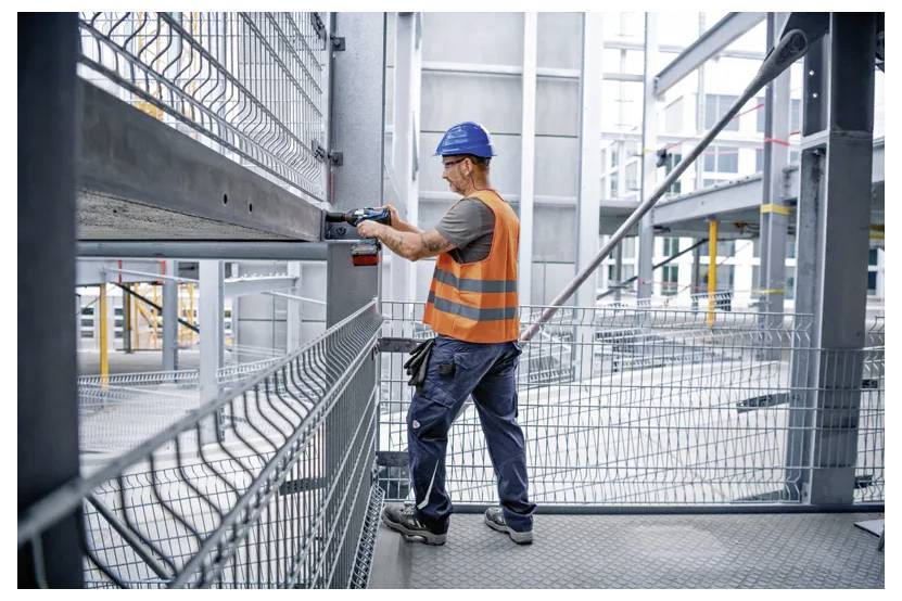 Ein Bauarbeiter mit blauem Helm und orangefarbener Warnweste bedient ein Elektrowerkzeug an einem Metallrahmen auf einer Baustelle.