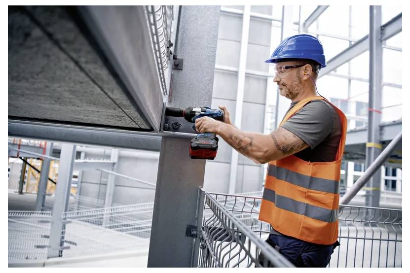 Ein Bauarbeiter mit blauem Schutzhelm und orangefarbener Warnweste benutzt einen Akkuschrauber an Metallträgern im Inneren eines sich im Bau befindenden Gebäudes.