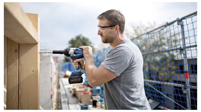 Ein Mann mit Sicherheitsbrille verwendet einen Akkuschrauber an einem Holzbalken auf einer Baustelle, umgeben von Sicherheitsabsperrungen und Bauausrüstung.