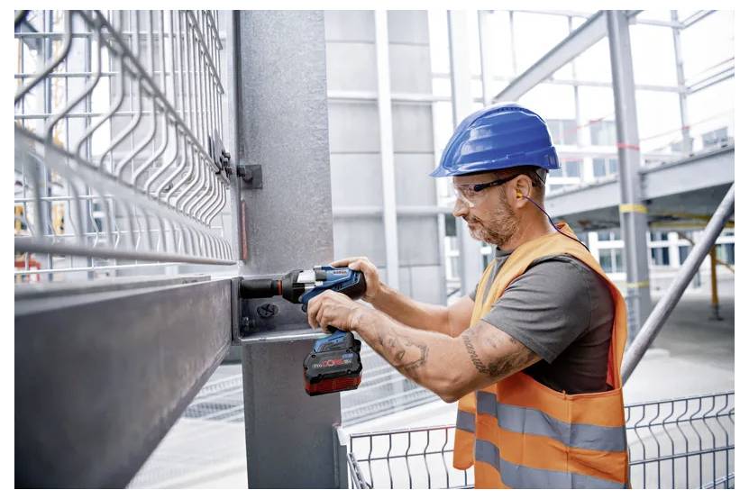 Ein Bauarbeiter mit blauem Helm und orangefarbener Warnweste verwendet ein Elektrowerkzeug, um einen Metallträger auf einer Baustelle zu befestigen.