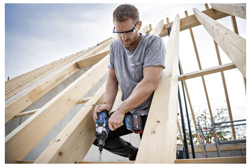 Eine Person mit Sicherheitsbrille bedient eine Bohrmaschine an Holzbalken eines sich im Bau befindlichen Daches vor einer teilweise bewölkten Himmelkulisse.
