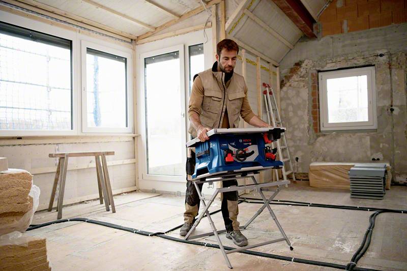 Person in Arbeitskleidung nutzt Werkzeug auf Baustelle. Unfertige Wände im Hintergrund, Baustelle wirkt aktiv und belebt.