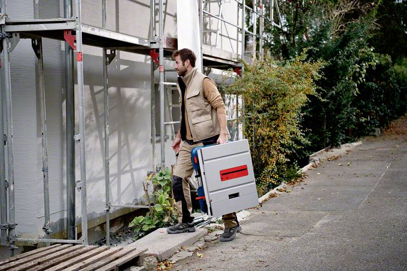 Eine Person in Arbeitskleidung trägt eine Werkzeugkiste auf einer Baustelle. Im Hintergrund sind Gerüste und Bäume zu sehen.