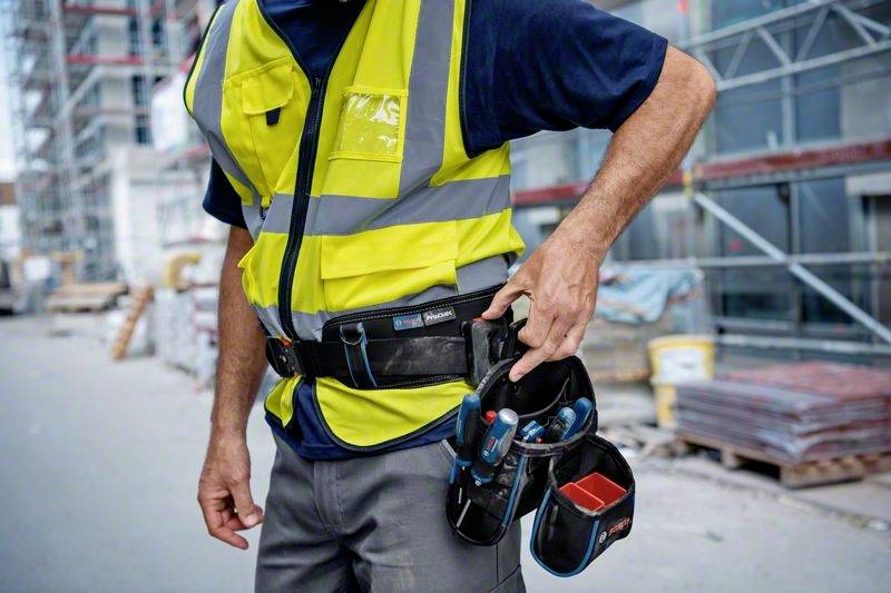Ein Bauarbeiter in gelber Warnweste steht auf einer Baustelle. Er trägt ein Werkzeugholster an der Hüfte und hält Werkzeug in der Hand.
