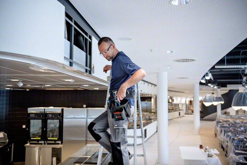 Ein Techniker arbeitet auf einer Leiter, um etwas in einem modernen, offenen Raum zu reparieren. Im Hintergrund sind Küchengeräte zu sehen.