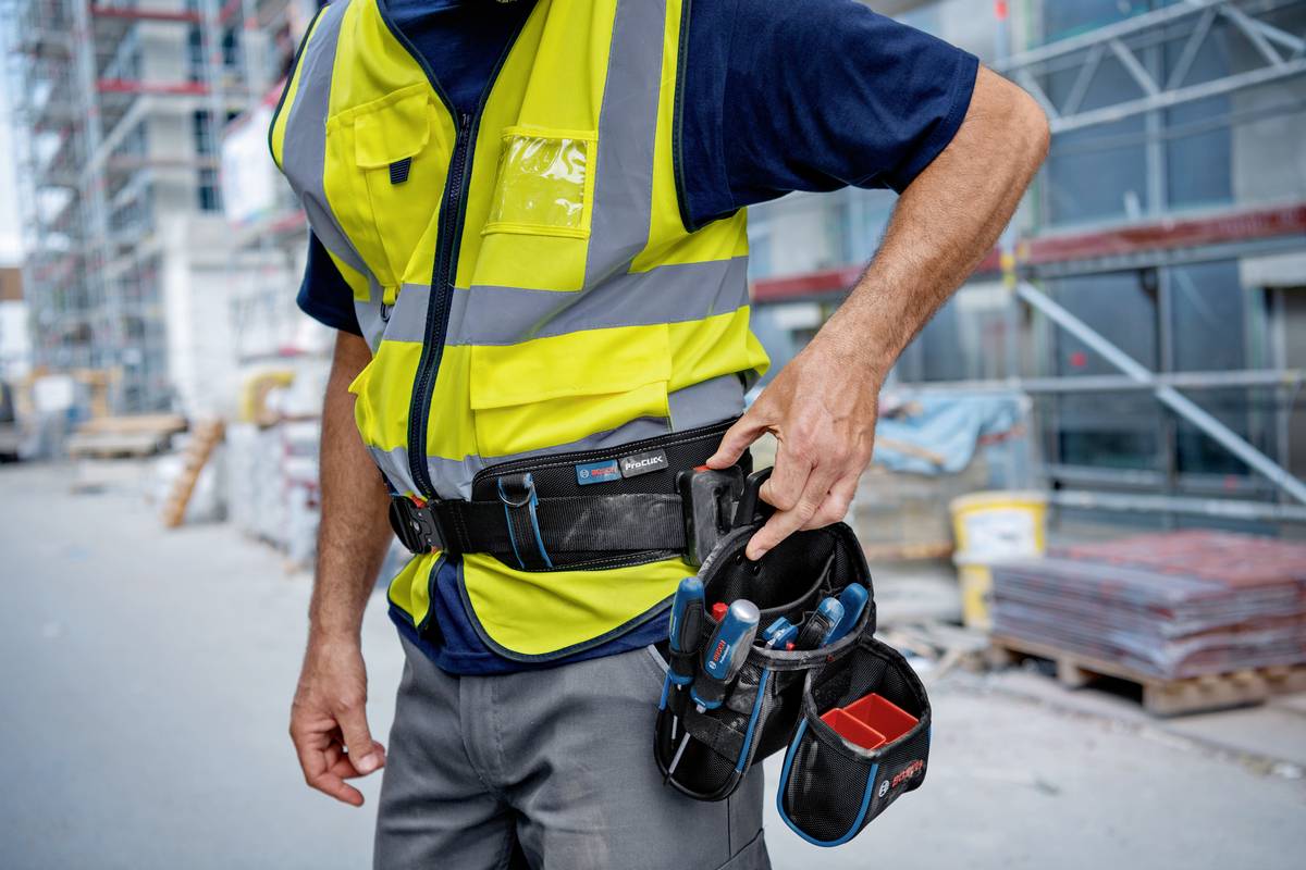 Eine Person auf einer Baustelle trägt eine gelbe Warnweste und einen Werkzeuggürtel mit mehreren Taschen. Hintergrund zeigt Gerüste.
