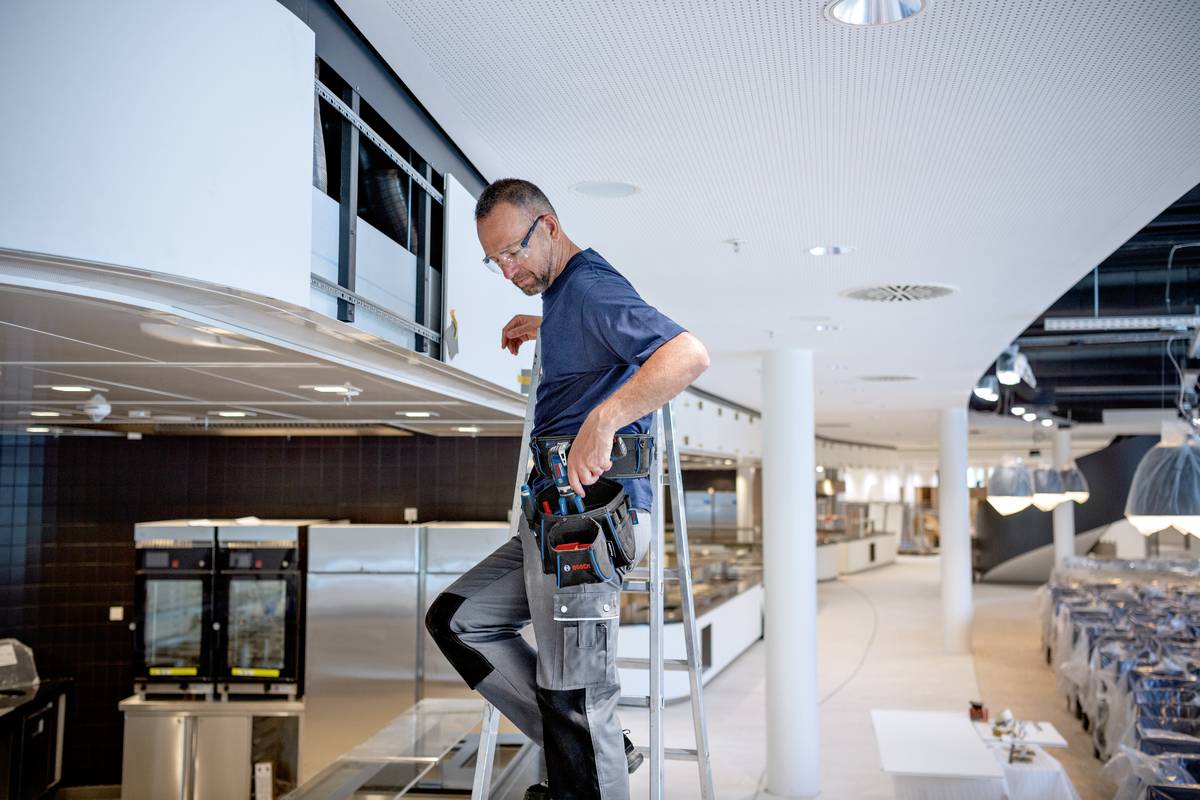 Ein Handwerker auf einer Leiter repariert eine Deckenleuchte in einem modernen Restaurant mit vielen Sitzbereichen.