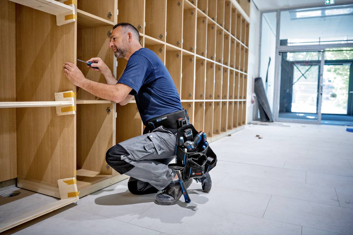 Ein Handwerker in blauer Kleidung montiert Regale in einem Raum. Er kniet auf dem Boden und hält ein Werkzeug in der Hand.