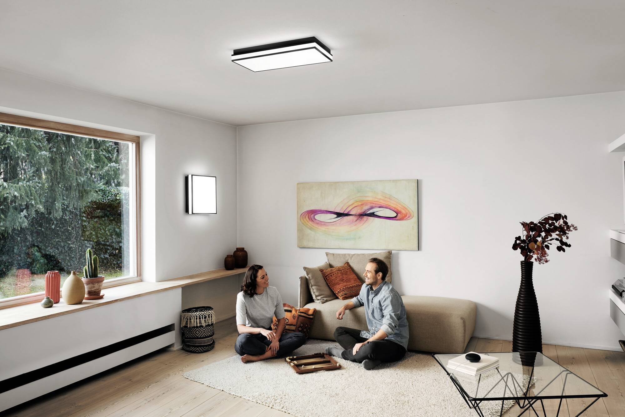 Ein Paar sitzt entspannt auf dem Boden in einem hellen Wohnzimmer, umgeben von Pflanzen und moderner Dekoration.