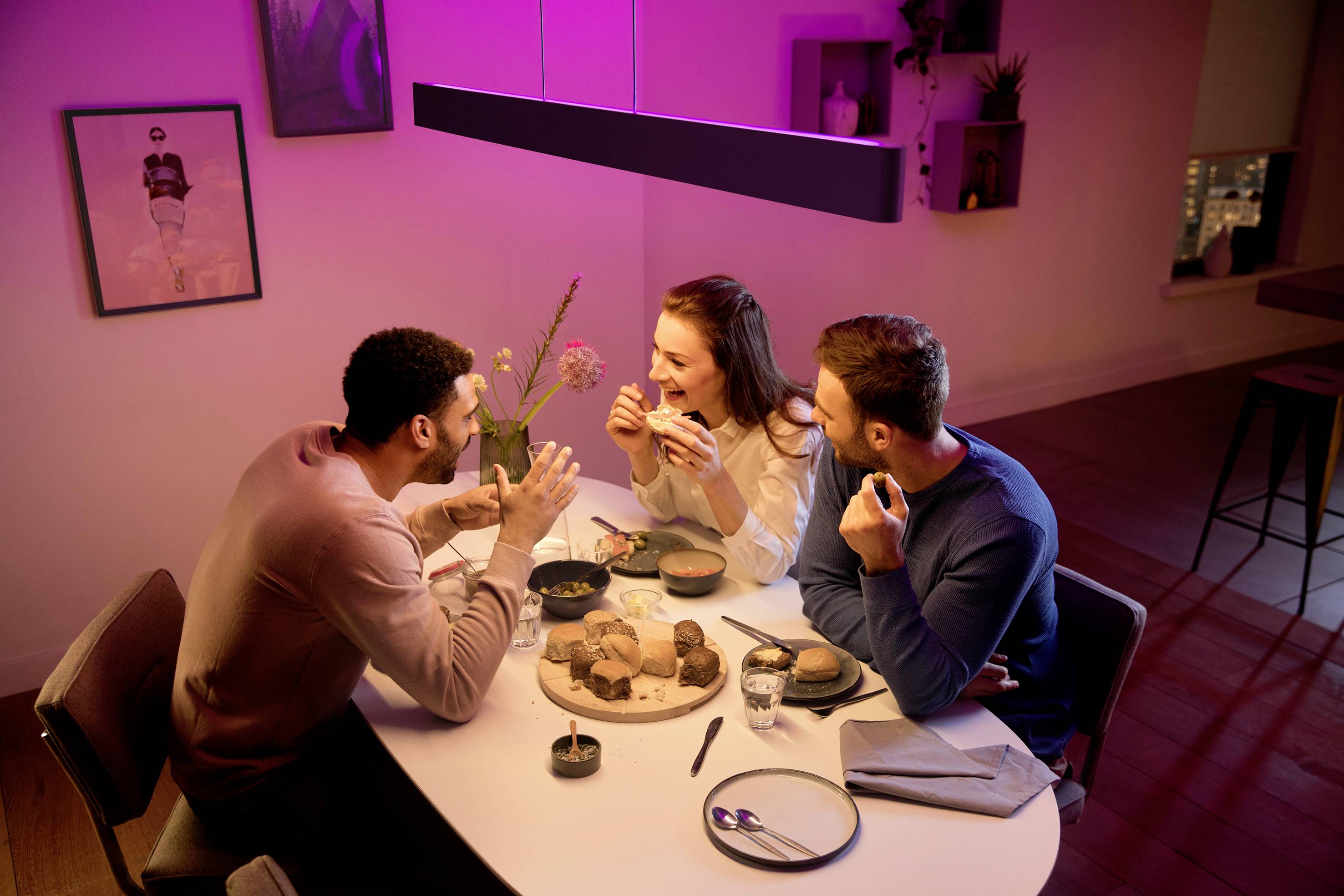Drei Personen sitzen an einem Tisch in einer gemütlichen Wohnung und unterhalten sich fröhlich beim Essen. Eine warme Beleuchtung schafft eine einladende Atmosphäre.