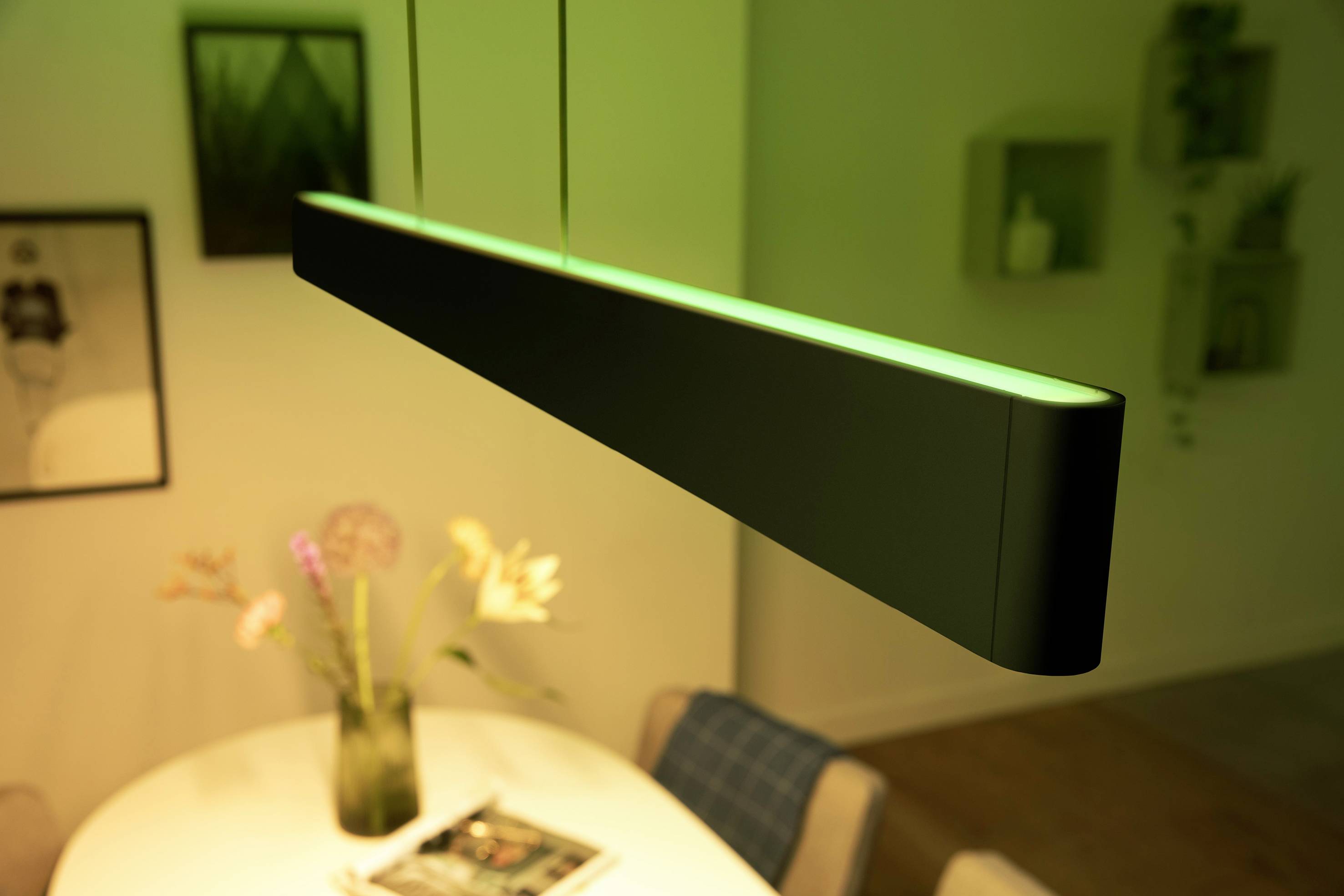 Hängelampe mit grünem Licht über rundem Tisch. Im Hintergrund sind unscharfe Bilderrahmen und Regale an der Wand zu sehen.