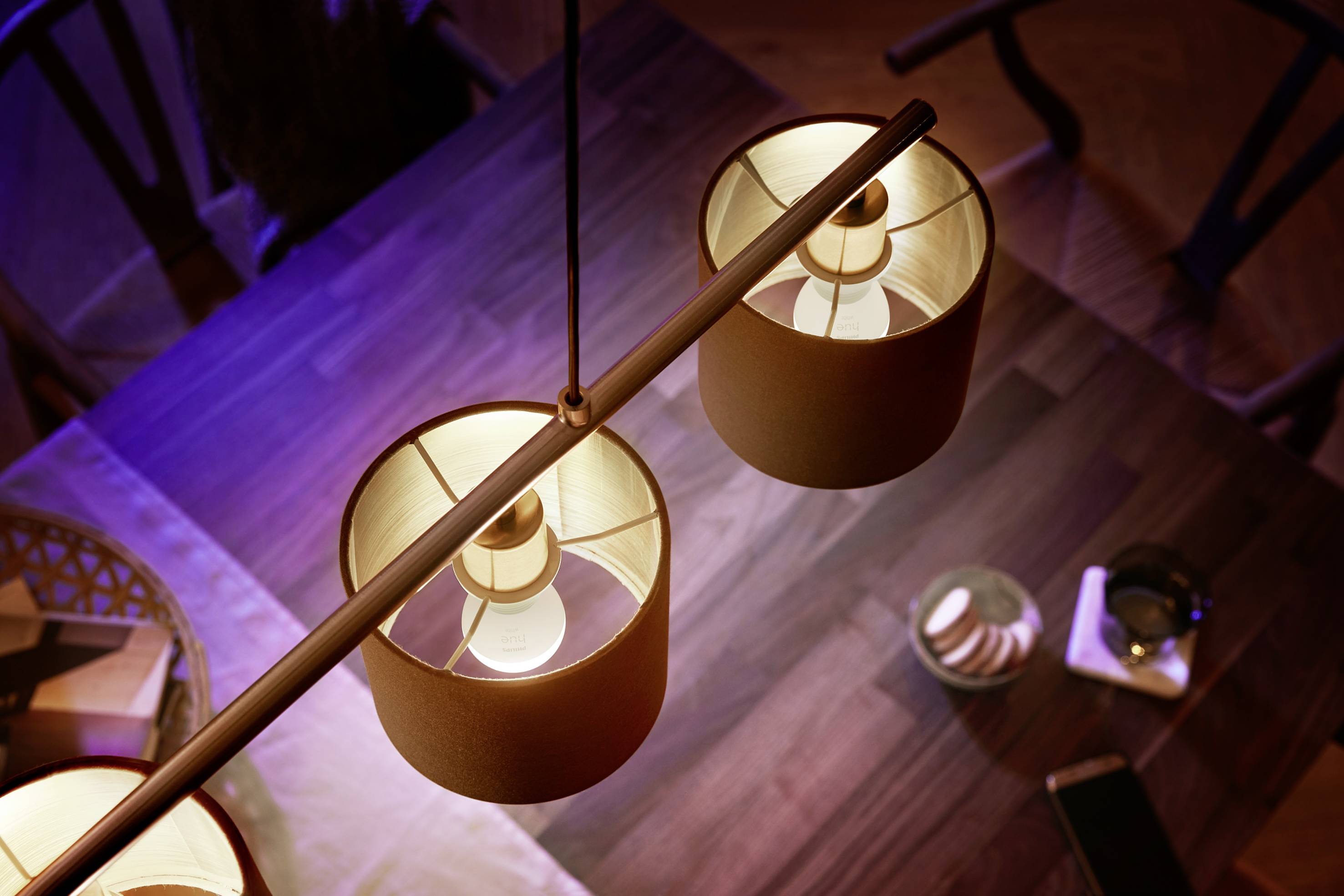 Zwei hängende Lampen leuchten über einem Esstisch aus Holz. Der Tisch hat Kerzen und ein Mobiltelefon. Stühle umgeben den Tisch.