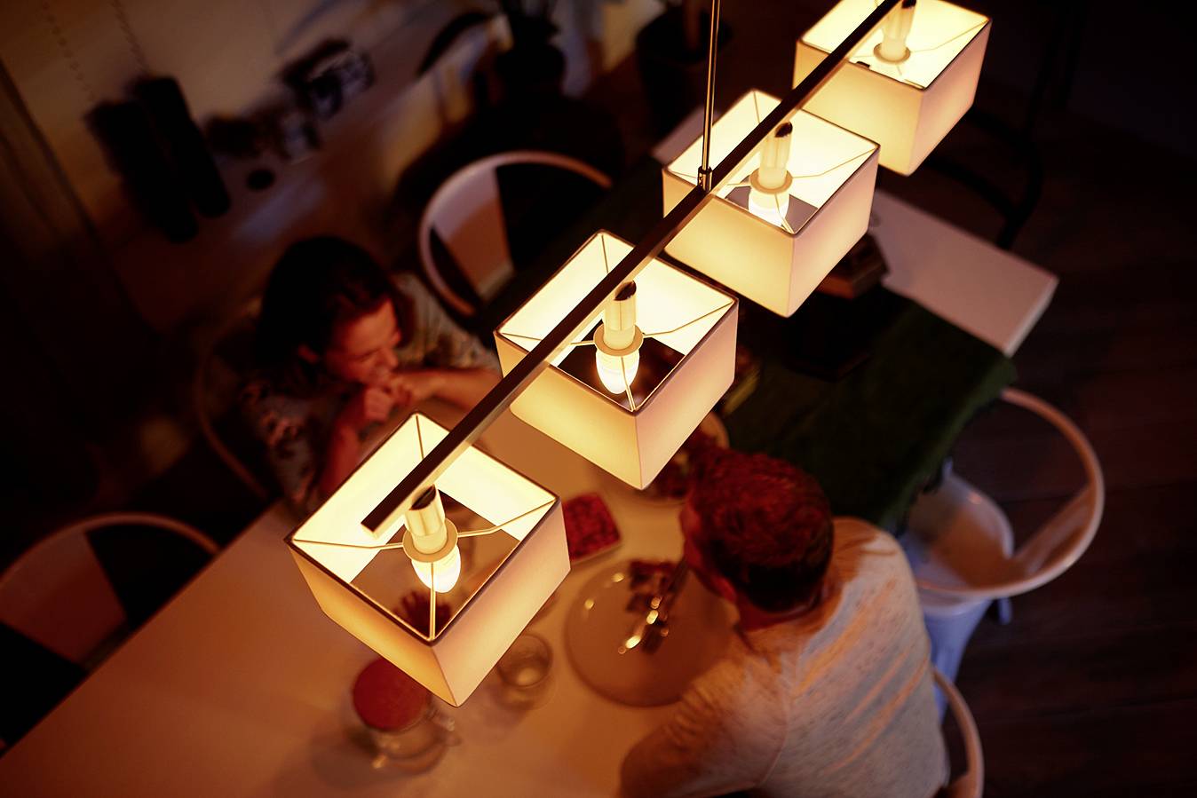 Vogelperspektive auf einen Esstisch mit zwei Personen, die unter einer Reihe von leuchtenden, modernen Lampen sitzen und essen.