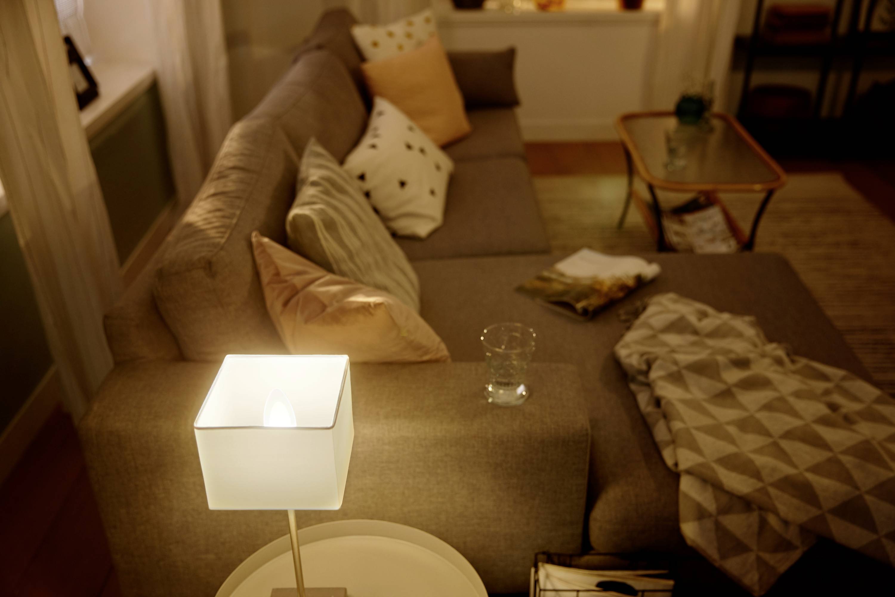 Wohnzimmer mit grauem Sofa und Kissen, auf dem ein Glas Wasser und eine Decke liegen. Eine Lampe leuchtet auf einem kleinen Tisch.
