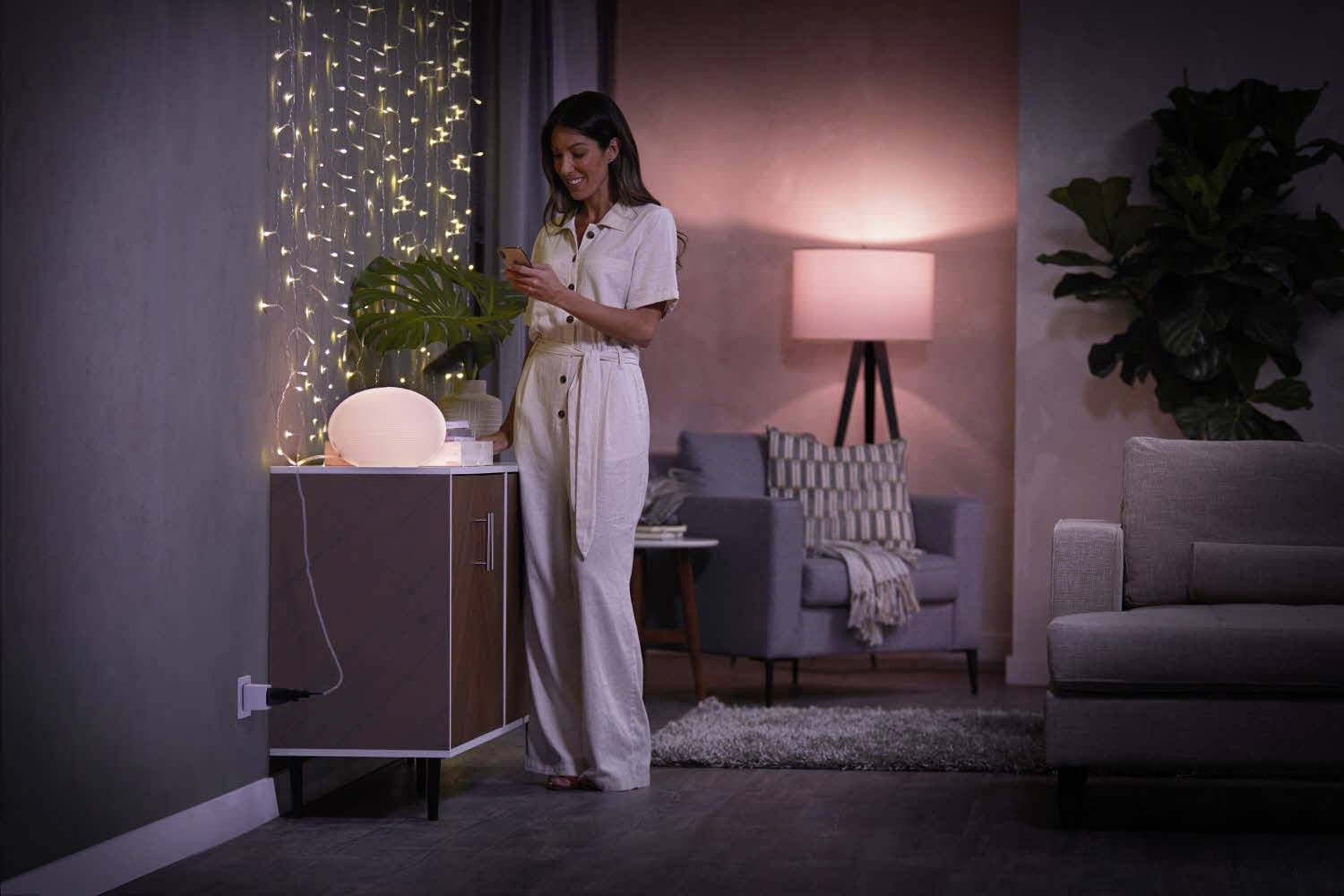 Eine Frau in einem gemütlich beleuchteten Wohnzimmer steht neben einem Tisch mit einer dekorativen Lampe und Lichterketten, lächelnd.