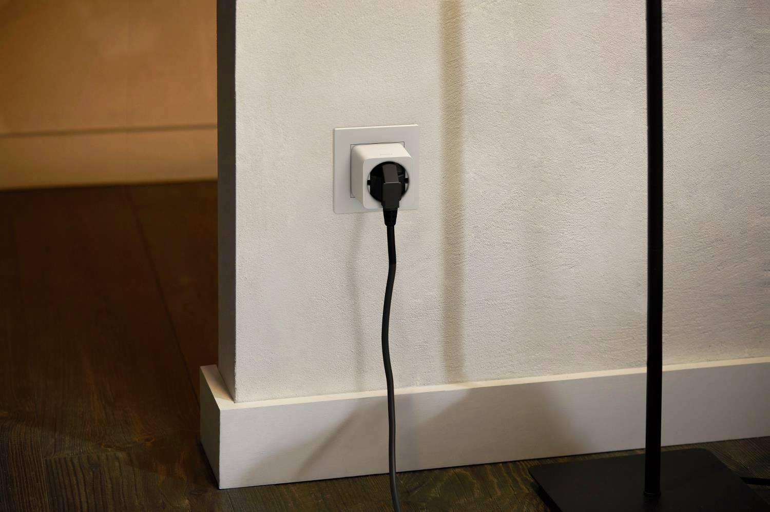 Eine schwarze Steckdose ist an einer weißen Wand montiert, ein Kabel ist eingesteckt und führt zum Boden. Links ist ein Holzboden sichtbar.