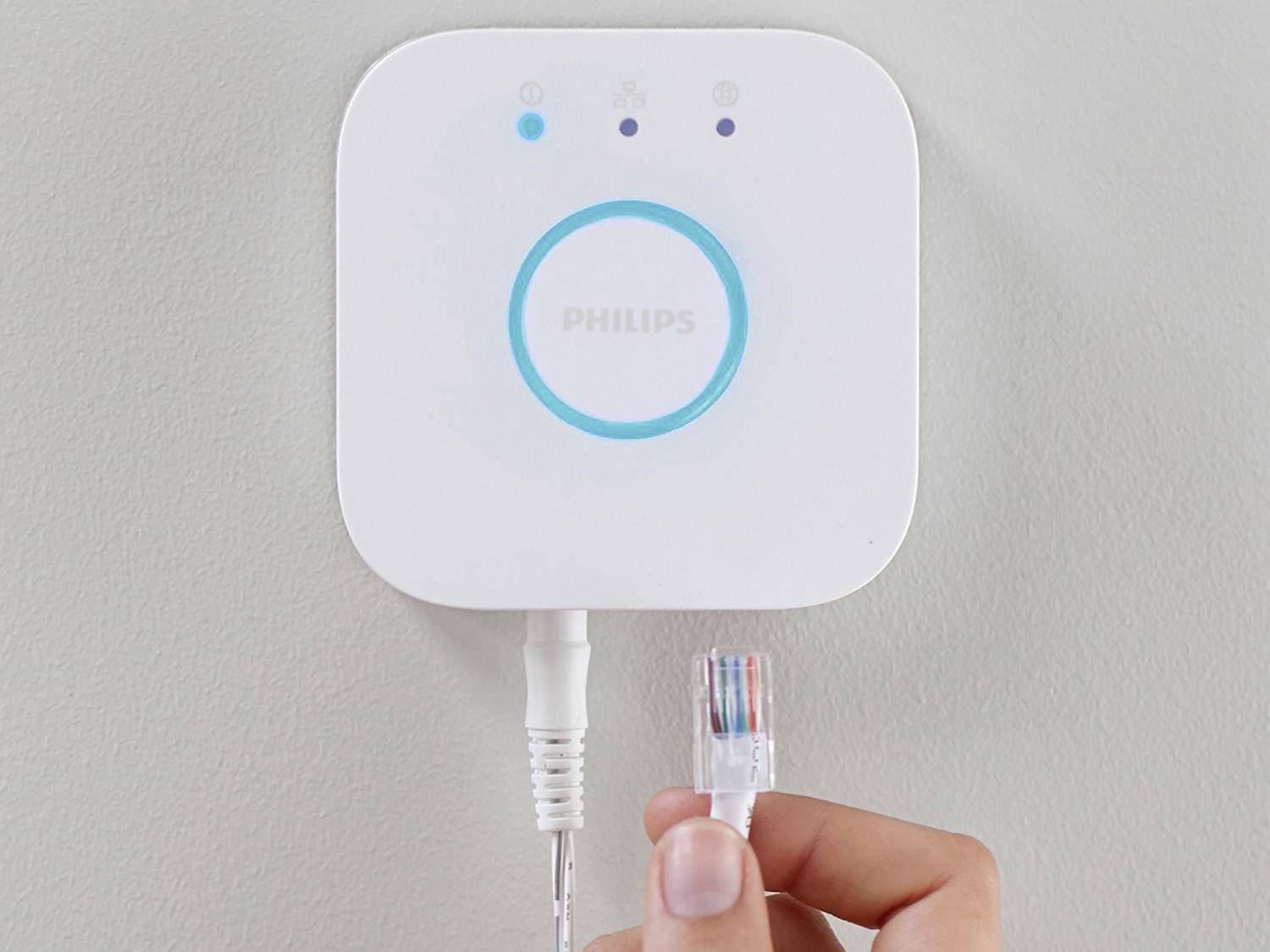 Eine Person hält einen Netzwerkstecker nahe an eine smart home Bridge mit leuchtendem blauen Ring und dem Logo 'Philips'.