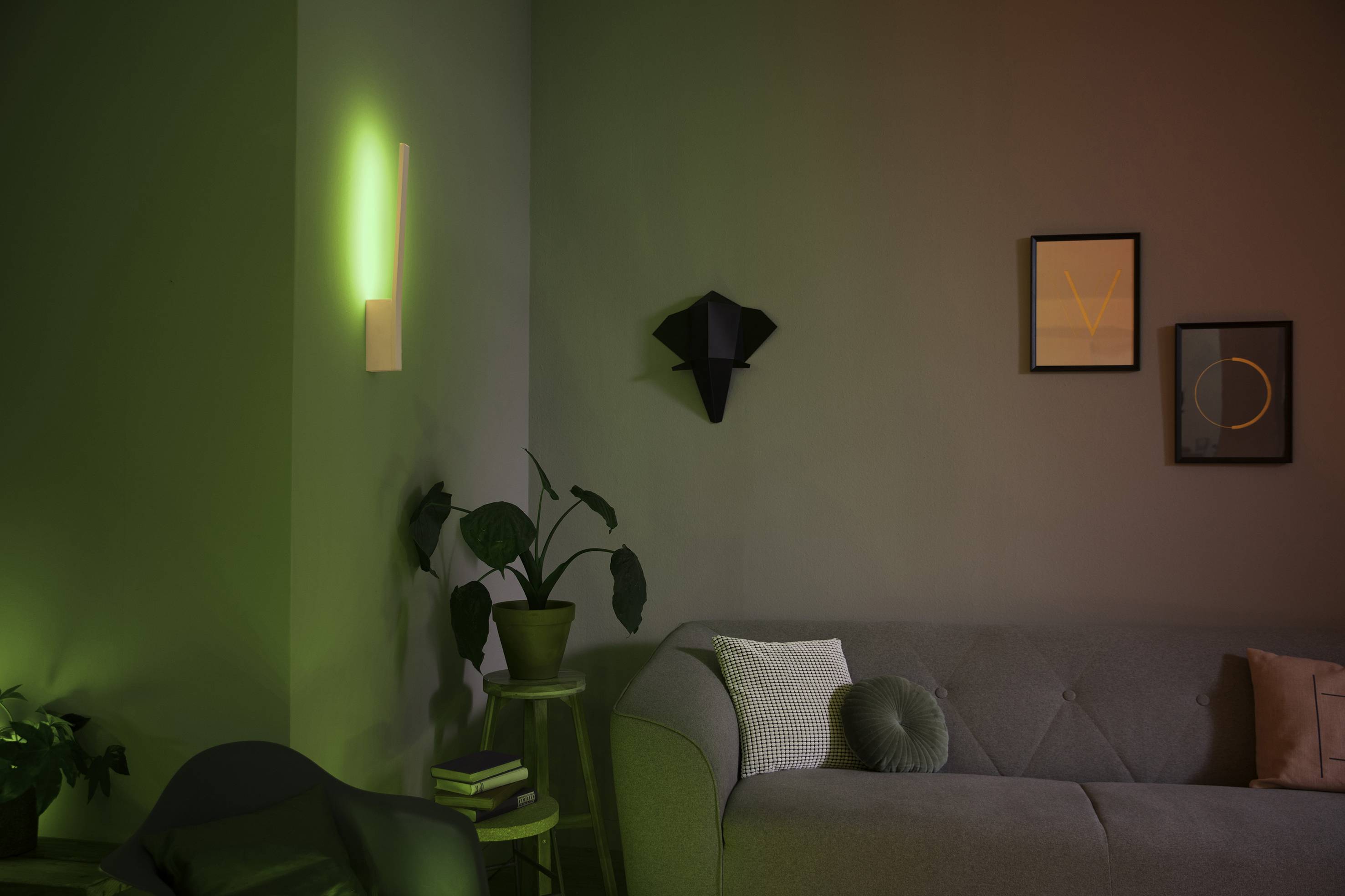 Ein Wohnzimmer mit grauem Sofa, grüner Pflanze, zwei Wandbildern und einer grünen Wandleuchte, die stimmungsvolles Licht erzeugt.