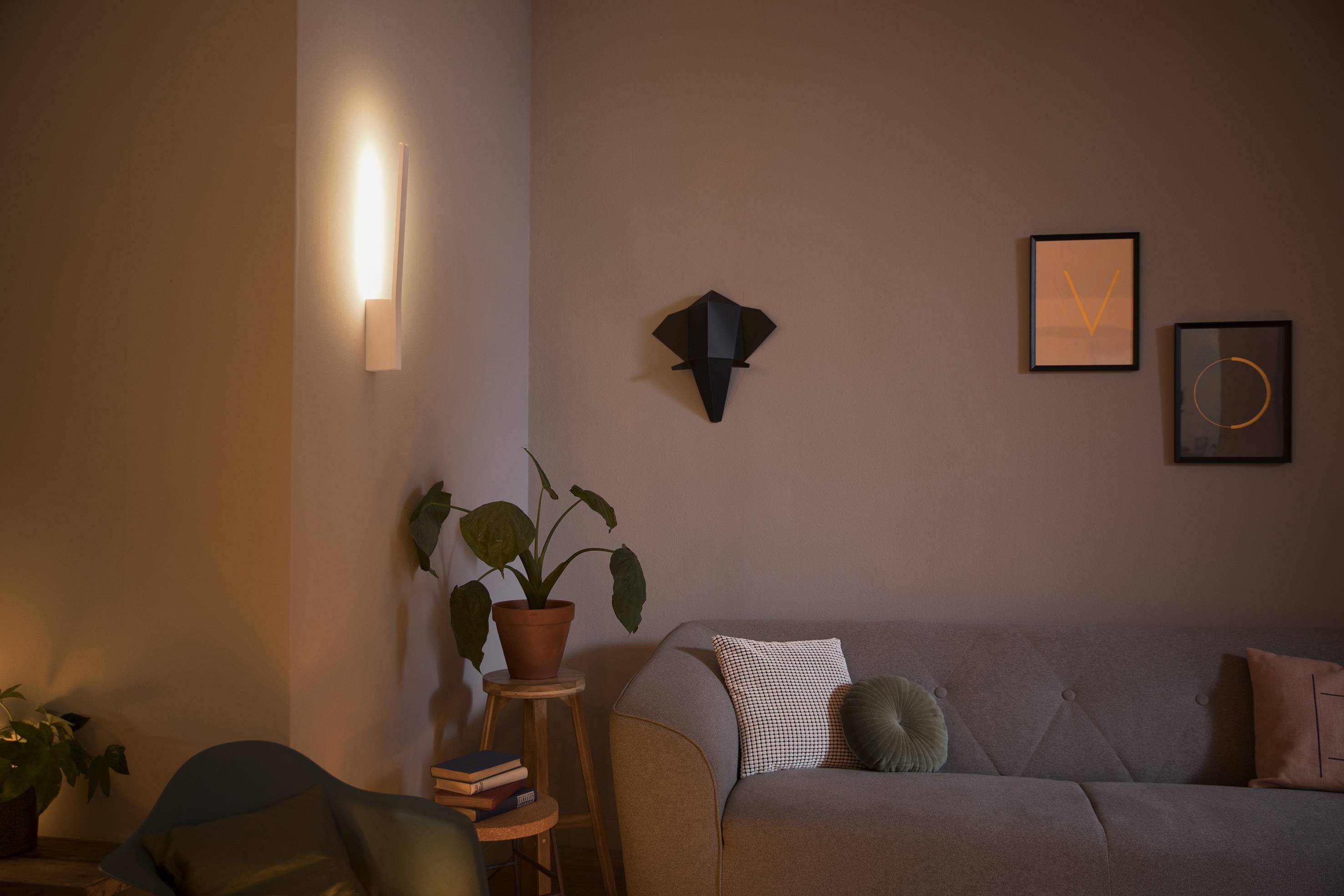 Wohnzimmer mit grauem Sofa, grünem Sessel, Pflanze auf einem Tisch und moderner Wandbeleuchtung; Bilderrahmen an der Wand.