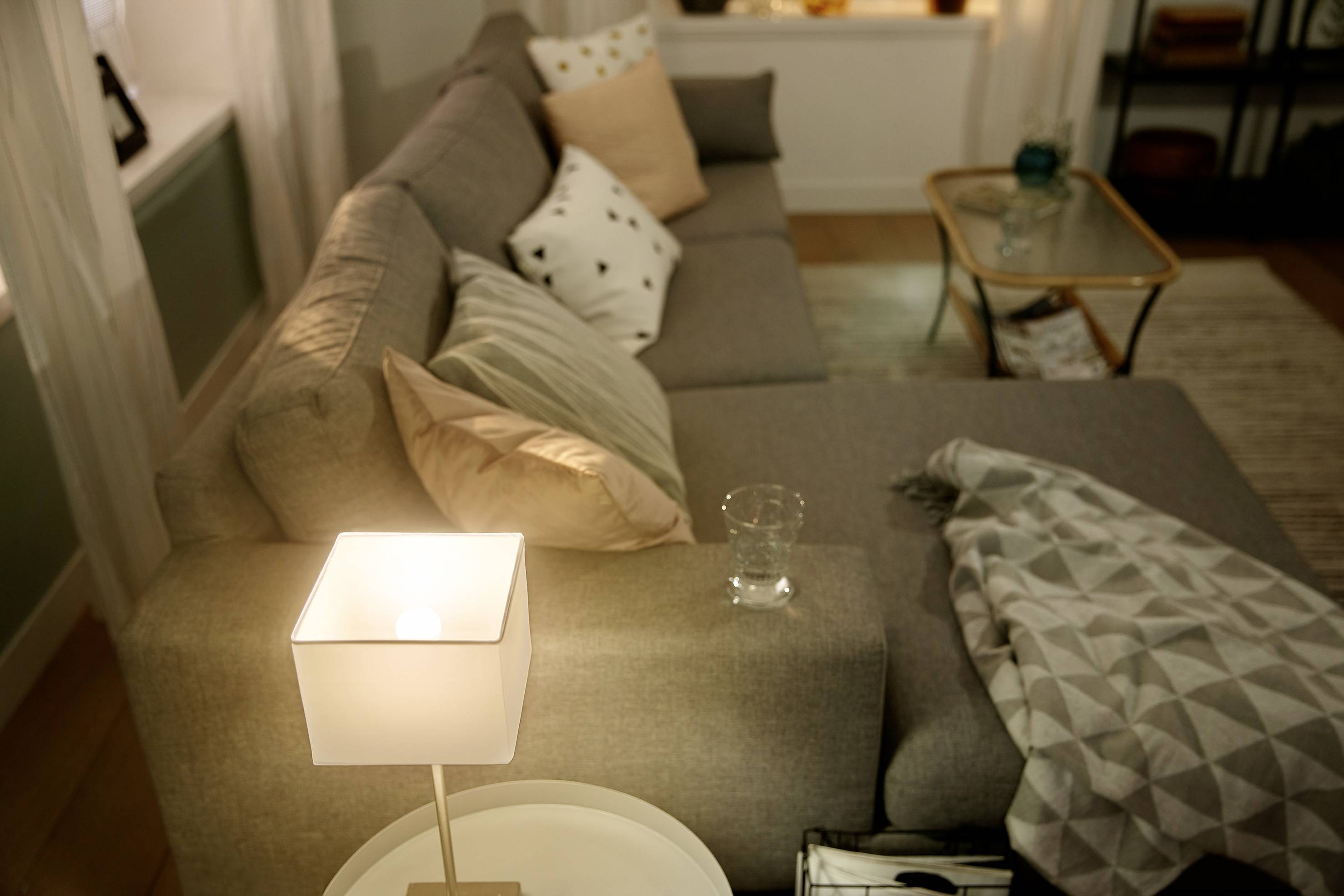 Wohnzimmer mit grauem Sofa, dekorativen Kissen, heller Tischlampe und kleinem Glastisch. Gemütliche, einladende Atmosphäre.