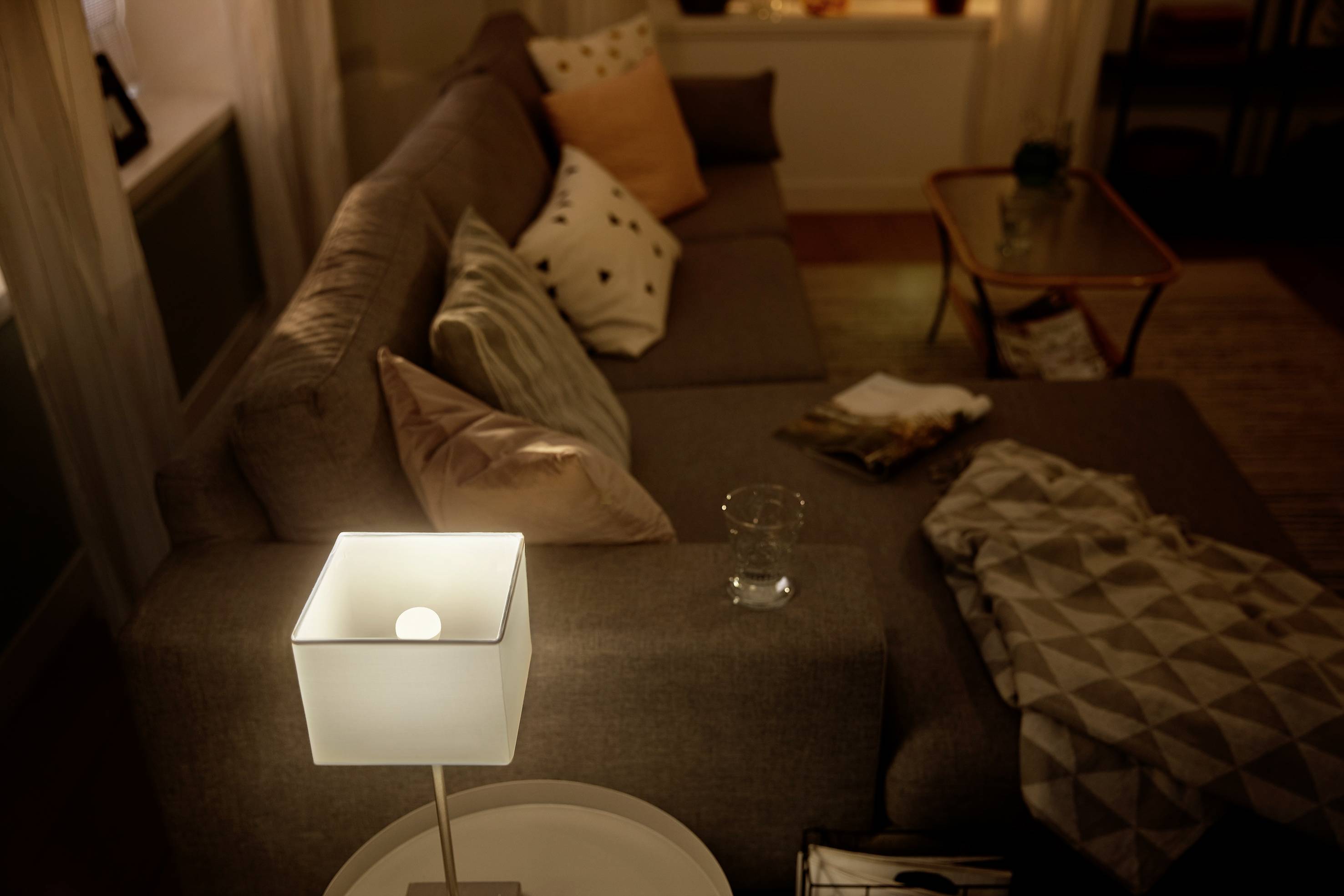 Gemütliches Wohnzimmer bei Dämmerung, Sofa mit Kissen, eine leuchtende Lampe auf einem Tisch, Plaids und Dekorelemente.