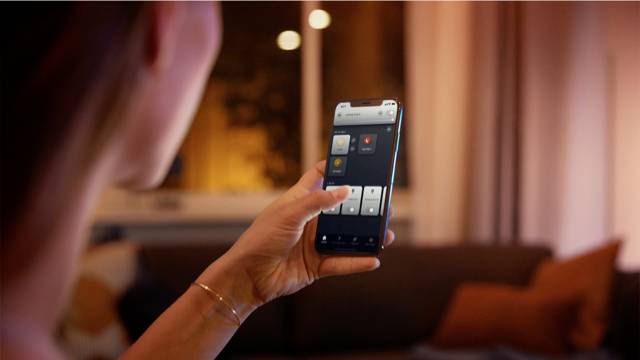 Eine Person hält ein Smartphone in der Hand und steuert damit eine App zur Hausautomation, während sie im Wohnzimmer sitzt.