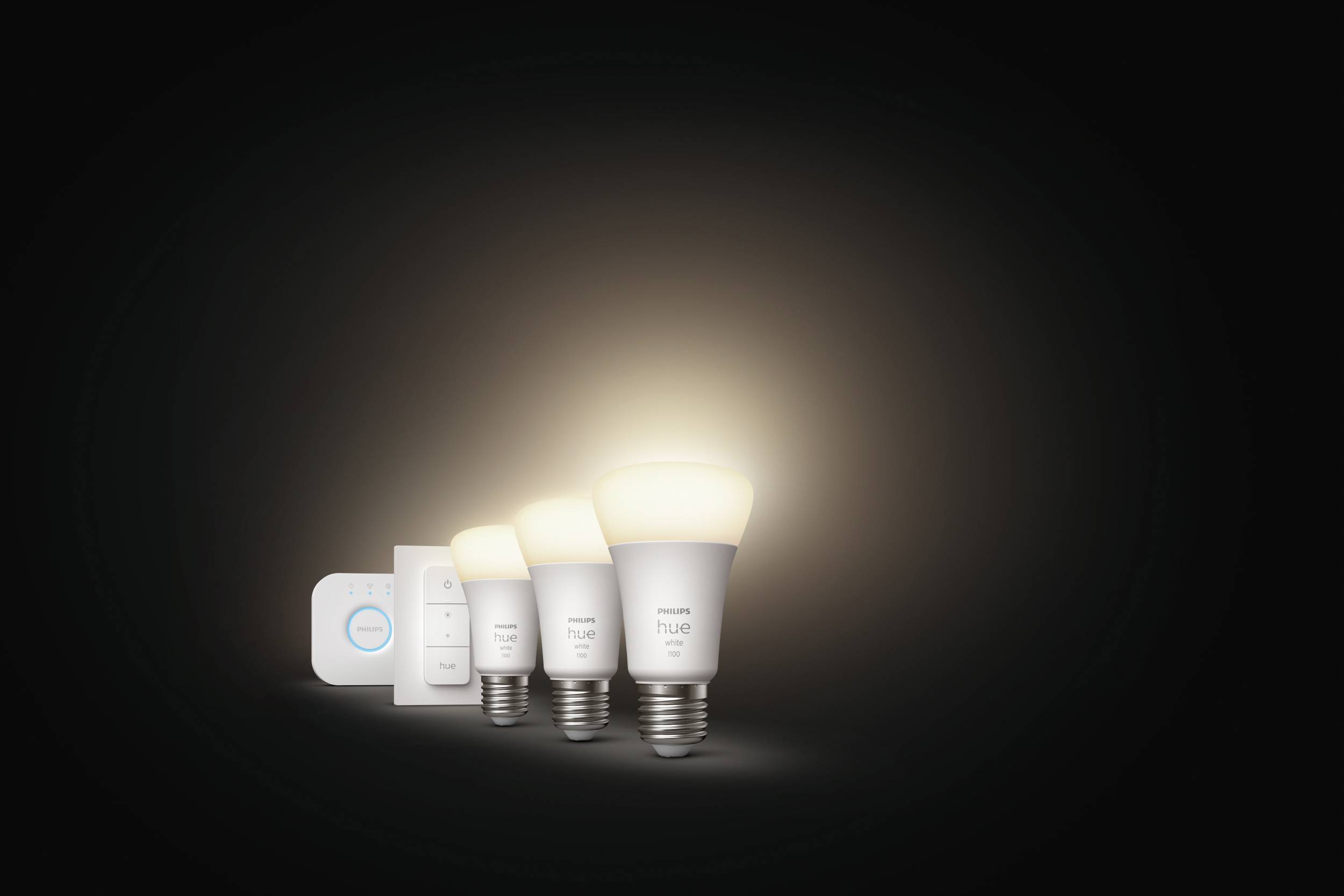 Drei Philips Hue Glühbirnen und ein Steuergerät vor dunklem Hintergrund. Die Glühbirnen leuchten sanft und zeigen ihre Farbvielfalt.
