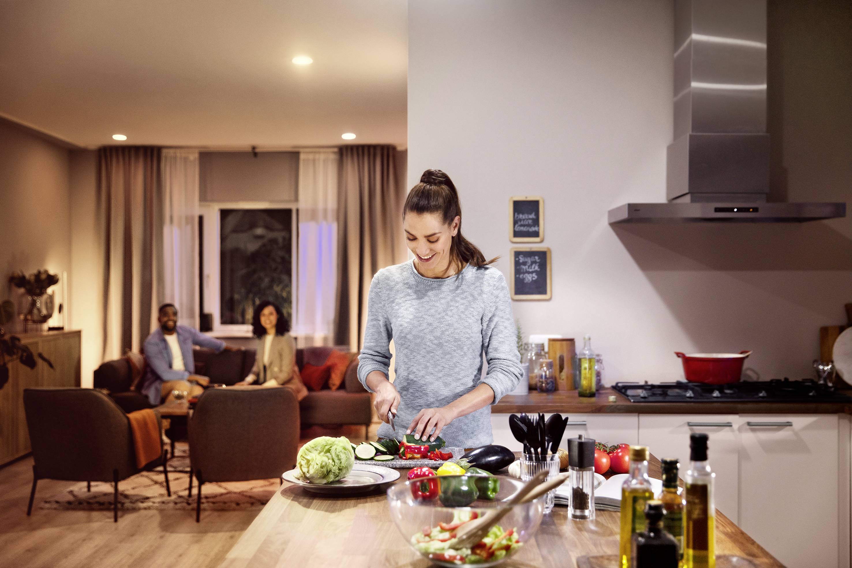 Eine Frau schneidet Gemüse in einer modernen Küche. Im Hintergrund sitzen zwei Personen auf dem Sofa und unterhalten sich.