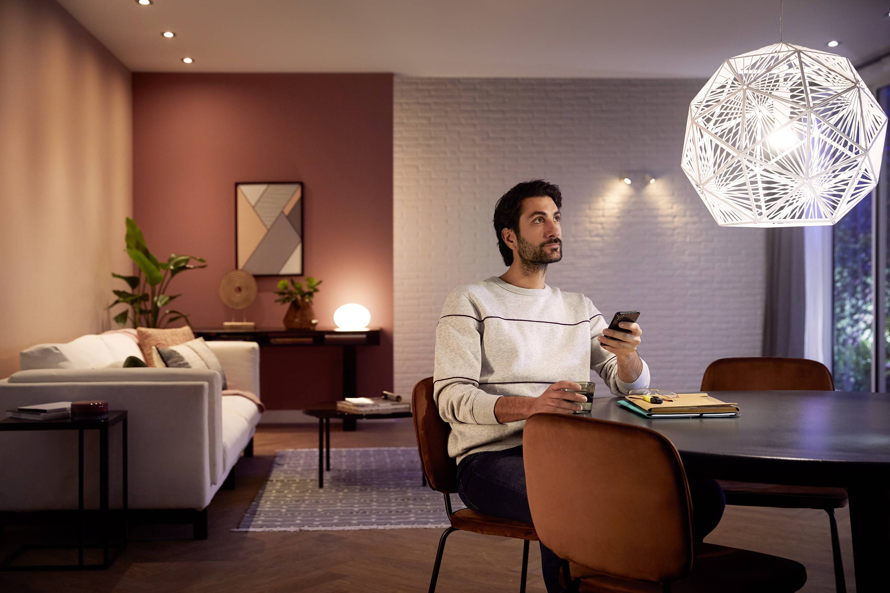 Ein Mann sitzt in einem Wohnzimmer an einem Tisch und hält eine Fernbedienung. Ein geometrisches Licht hängt über ihm.