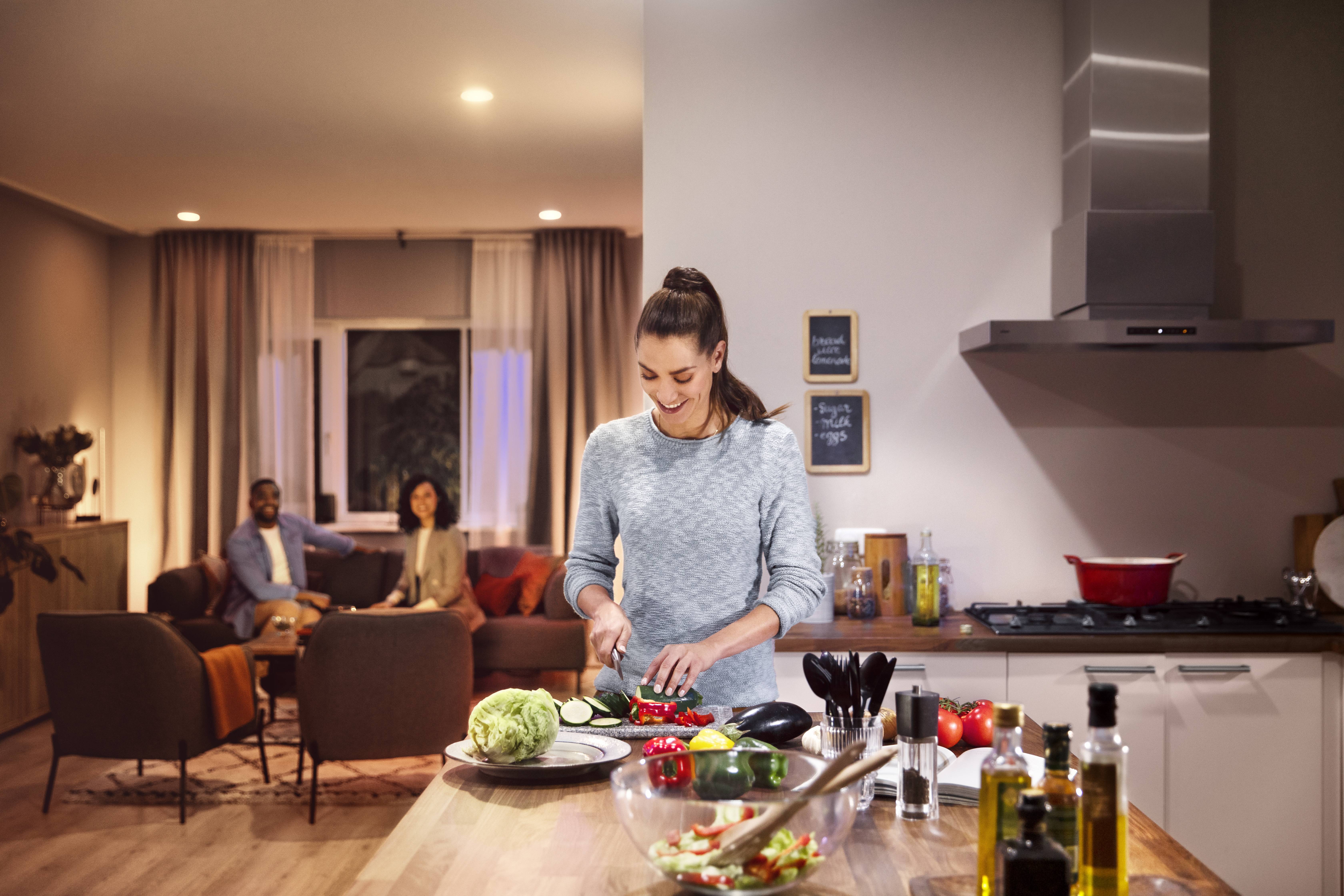 Eine Frau schneidet Gemüse in einer modernen Küche. Im Hintergrund sitzen zwei Personen im Wohnzimmer und unterhalten sich.