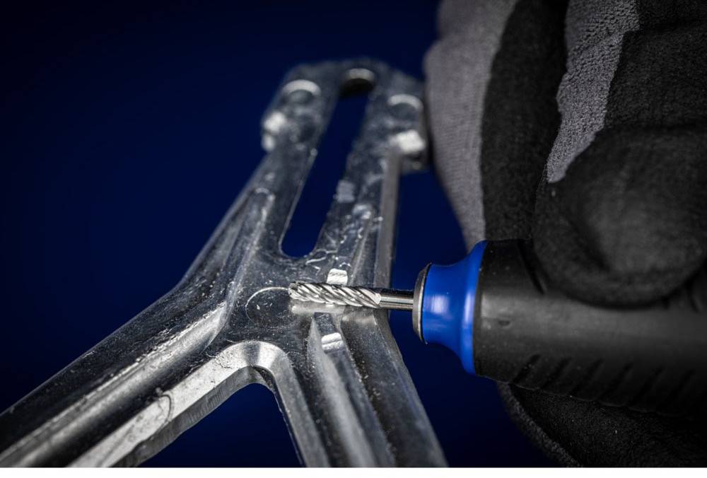 Eine Hand in einem Handschuh bearbeitet mit einem rotierenden Werkzeug ein Metallstück. Fokus auf Präzision und Detailarbeit.