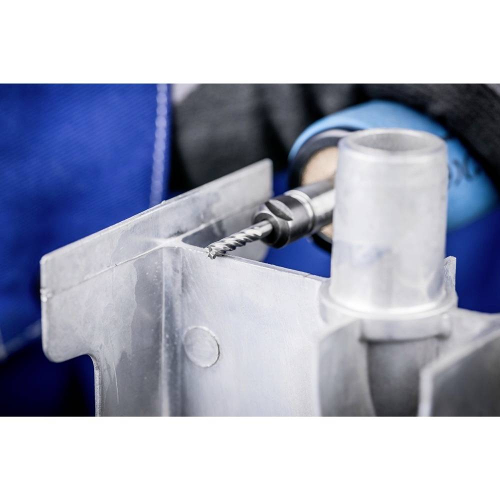 Eine Person, die mit einer Bohrmaschine ein Loch in ein Metallteil bohrt. Sie trägt Arbeitskleidung.