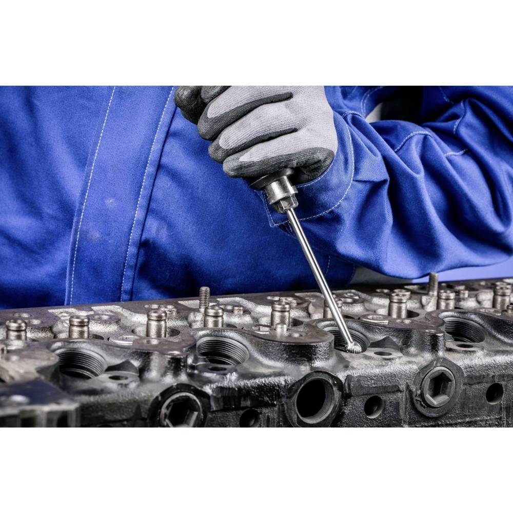 Ein Mechaniker in blauer Arbeitskleidung arbeitet mit einem Werkzeug an einem Motorteil, fokussiert auf die Präzision der Arbeit.