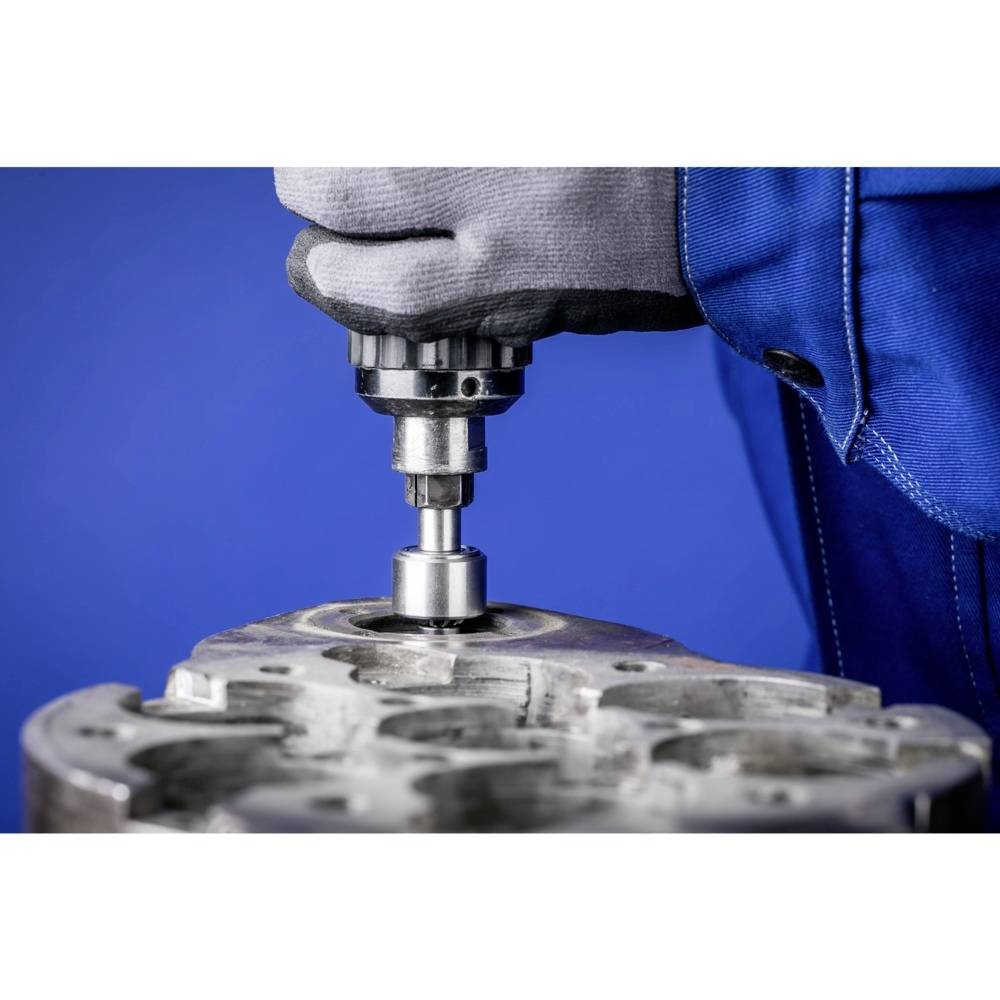 Ein Mechaniker bearbeitet eine Metalloberfläche mit einem Bohrer. Die Person trägt Arbeitshandschuhe und eine blaue Arbeitskleidung.