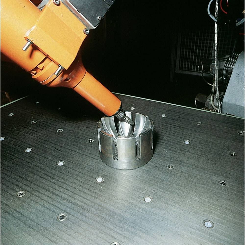 Ein orangefarbener Roboterarm bearbeitet ein zylindrisches Metallteil auf einem Arbeitstisch. Im Hintergrund ist eine Maschine zu sehen.