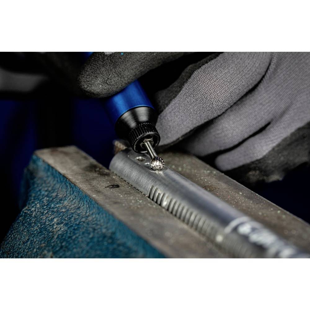 Nahaufnahme von Händen in grauen Handschuhen, die ein Metallstück mit einem blauen Werkzeug in einem Schraubstock bearbeiten.