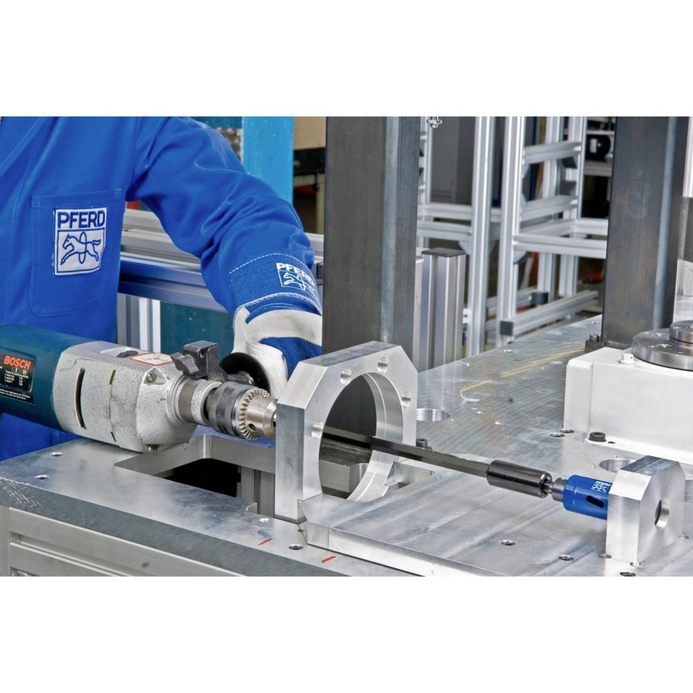 Eine Person in blauer Schutzkleidung bedient eine Bohrmaschine, die an eine Metallhalterung angeschlossen ist. Im Hintergrund sind Regale zu sehen.