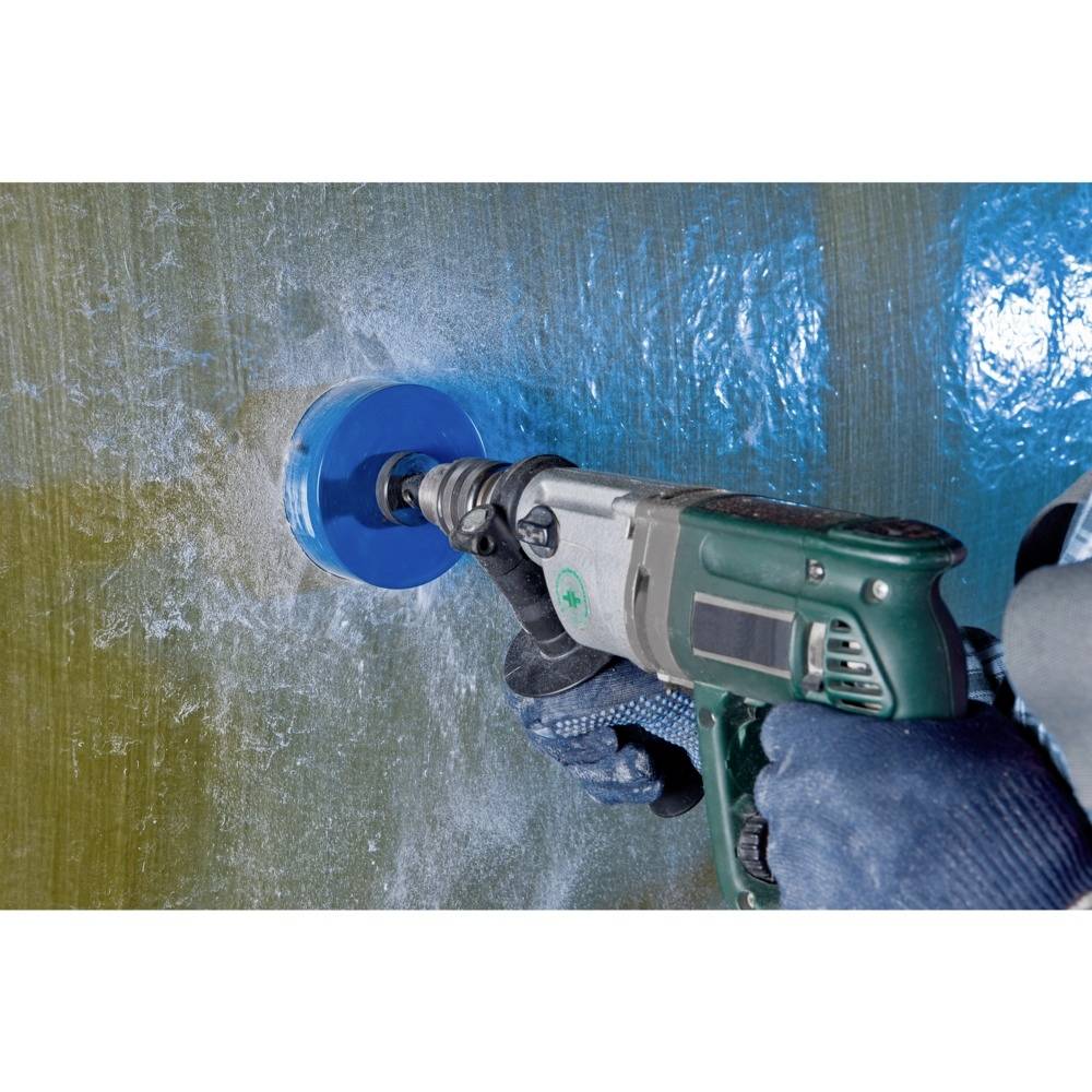 Eine Person benutzt eine elektrische Bohrmaschine, um ein Loch in eine blaue, raue Oberfläche zu bohren. Die Szene zeigt Handschuhe und einen blauen Bohreraufsatz während des Einsatzes.