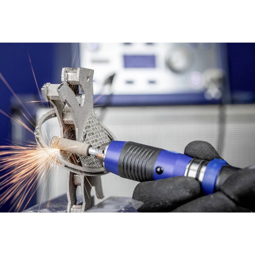 Eine gloved Hand bearbeitet eine komplexe Metallstruktur mit einem rotierenden Werkzeug, während Funken sprühen.