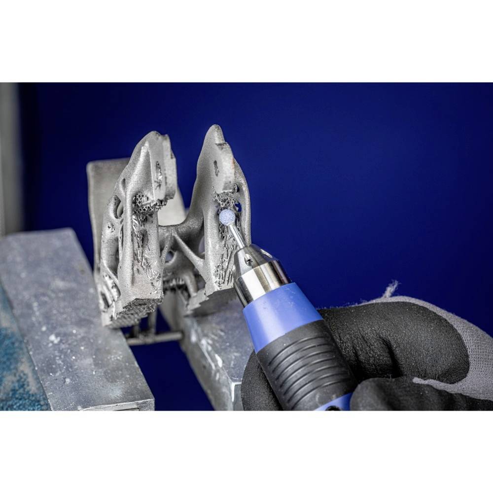 Eine Hand mit Handschuh hält ein elektrisches Werkzeug und bearbeitet ein graues, komplex geformtes Metallobjekt auf einer Werkbank.
