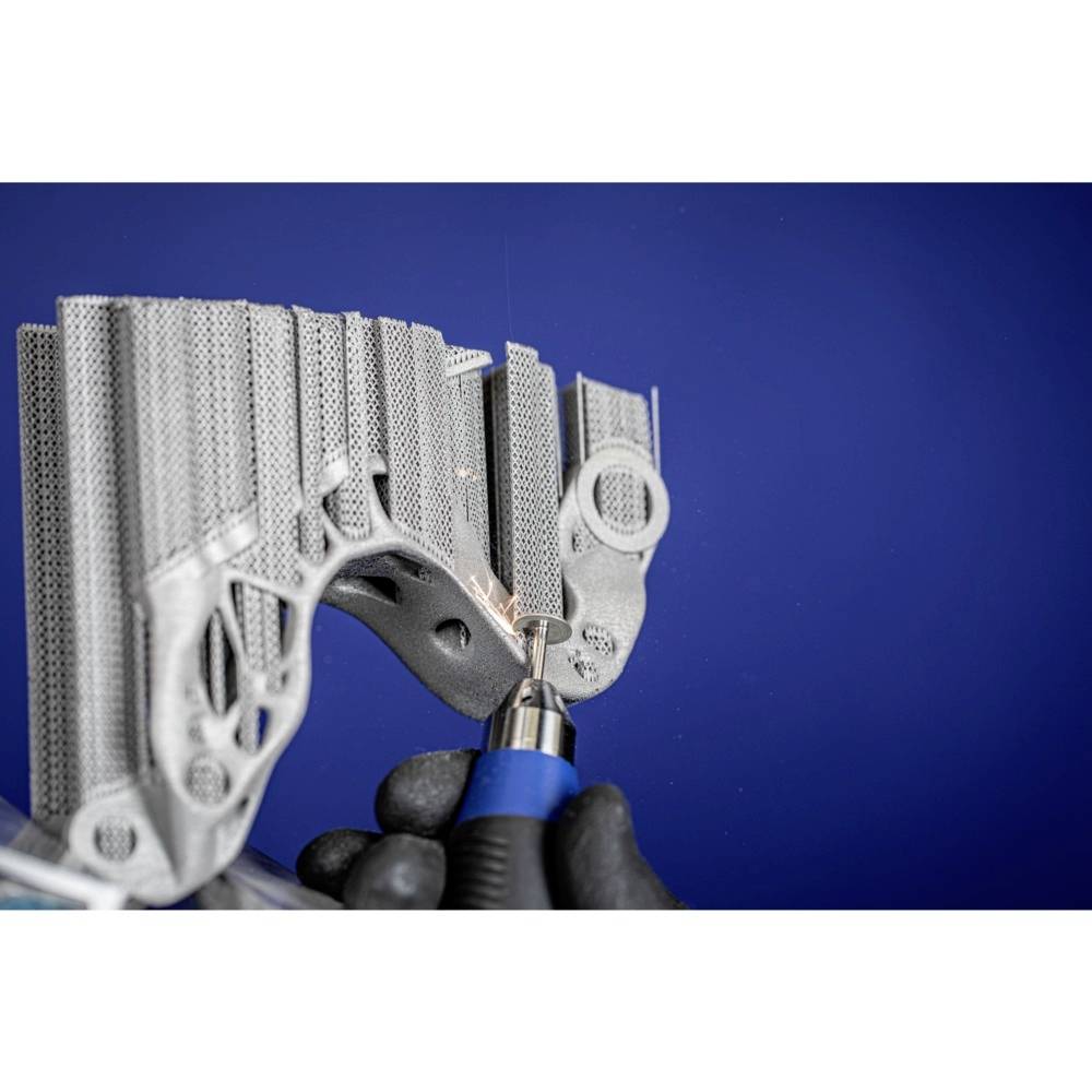 Eine Hand hält ein Werkzeug und bearbeitet ein komplexes, metallisches 3D-Druckteil vor blauem Hintergrund.