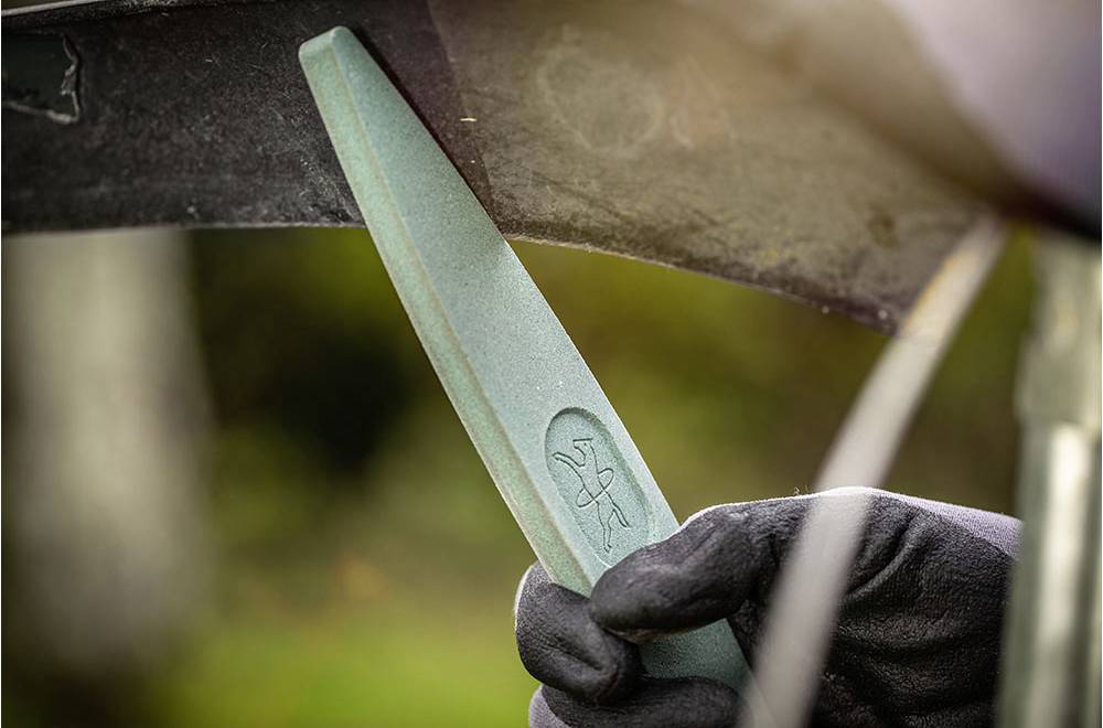 Eine behandschuhte Hand hält einen Schleifstein gegen eine Klinge, um sie zu schärfen. Im Hintergrund unscharfes Grün.