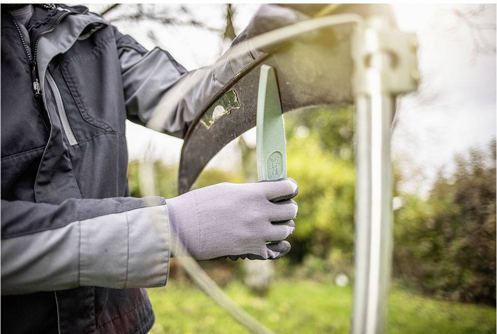 Person schärft eine Sense mit einem Schleifstein im Freien. Die Person trägt Handschuhe und eine Jacke.