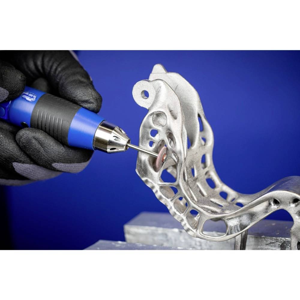 Hand hält ein rotierendes Werkzeug, das ein komplex geformtes Metallstück bearbeitet. Mehrere Öffnungen im Metall sind sichtbar.