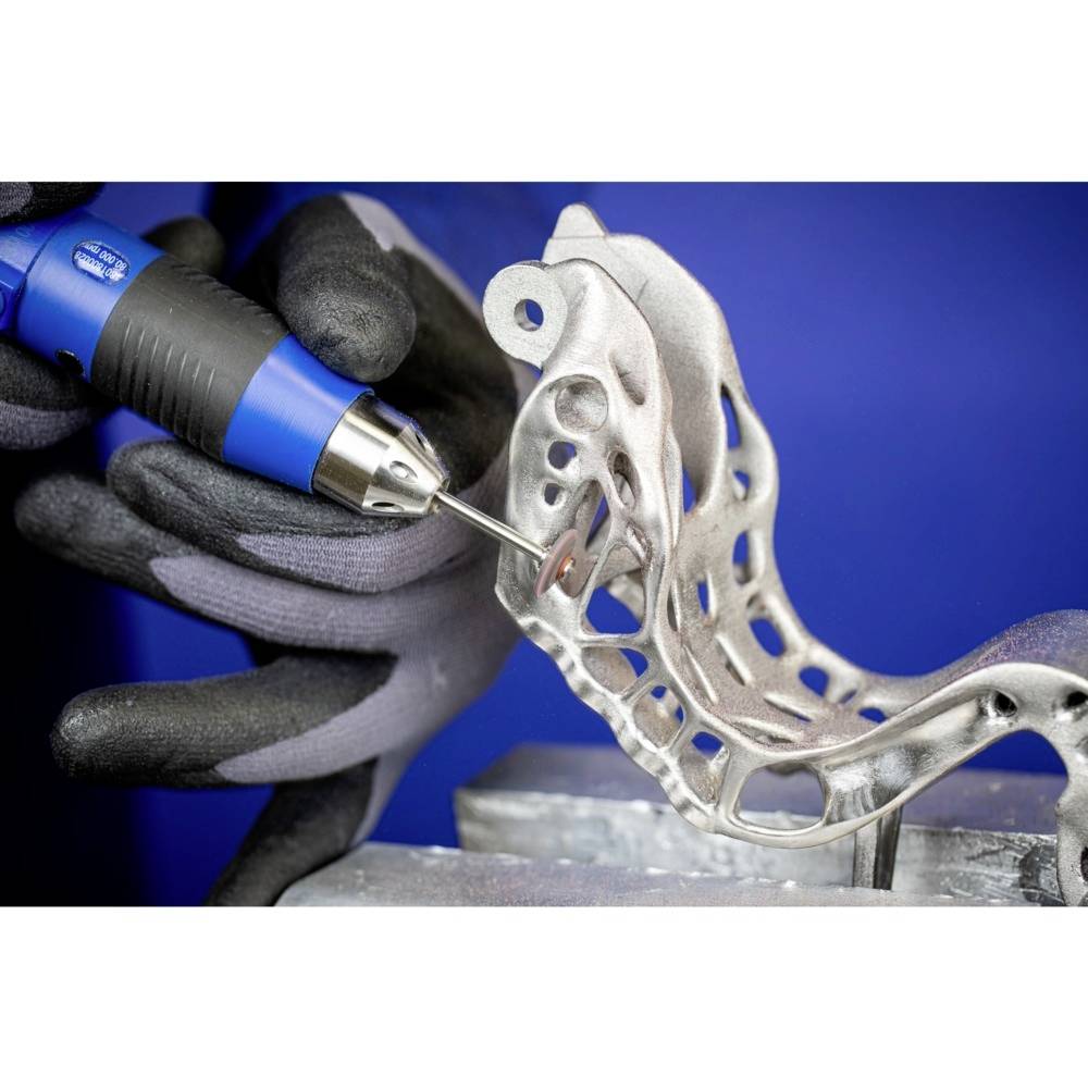 Eine Person bearbeitet ein komplexes Metallbauteil mit einem blauen Handwerkzeug. Das Bauteil hat viele Löcher und geschwungene Formen.