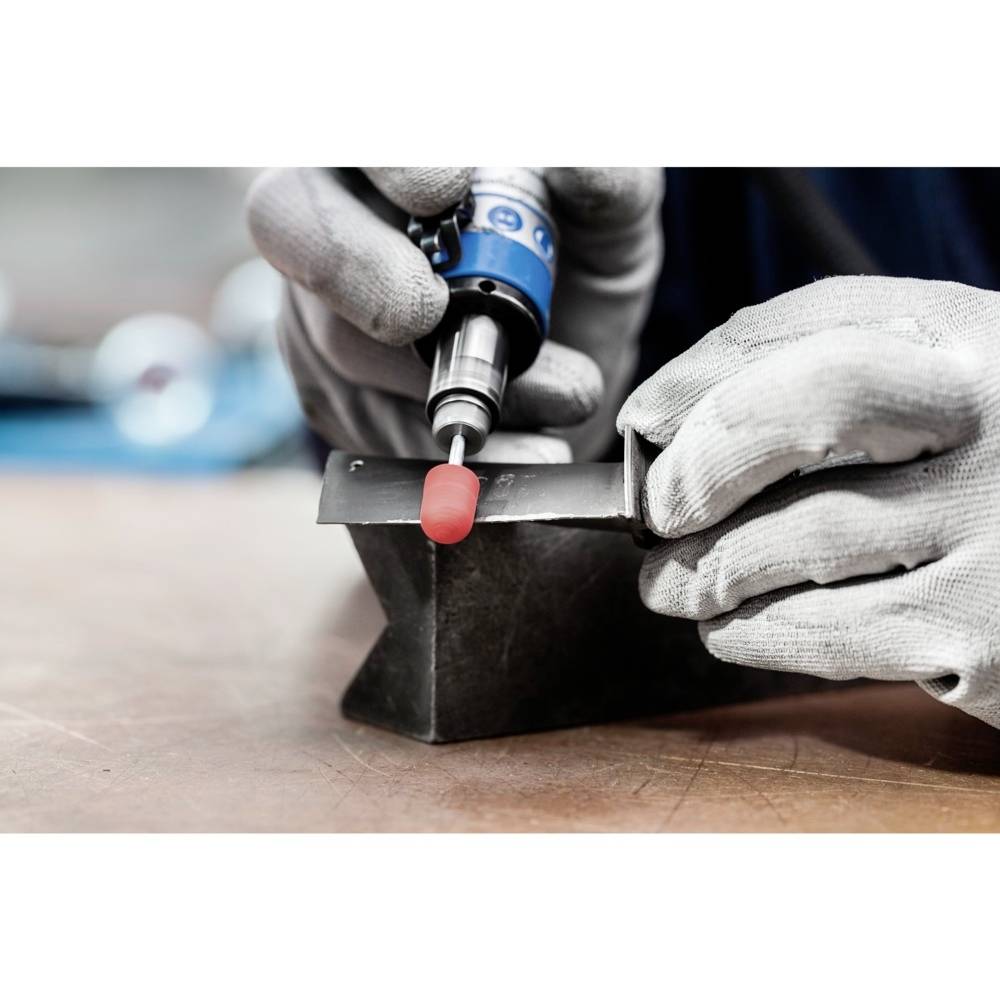 Eine Person mit Handschuhen schleift mit einem rotierenden Werkzeug einen Metallwinkel auf einem Tisch. Schwerpunkt auf Präzisionsarbeit.