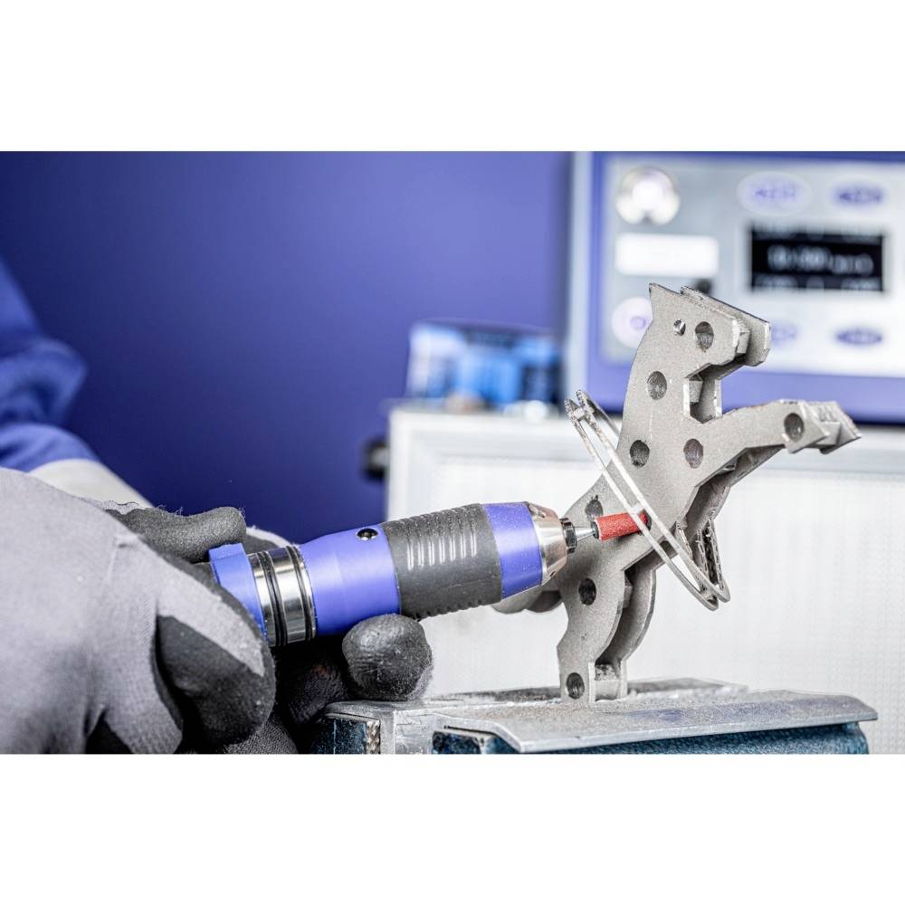 Eine Person in Handschuhen arbeitet mit einem blauen Werkzeug an einem metallischen Bauteil. Hintergrund unscharf mit technischen Geräten.