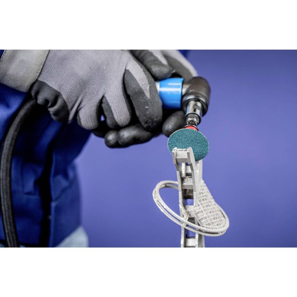 Handwerker schleift Metallstück mit Schleifwerkzeug. Er trägt graue Handschuhe und blaue Schutzkleidung. Hintergrund unscharf blau.