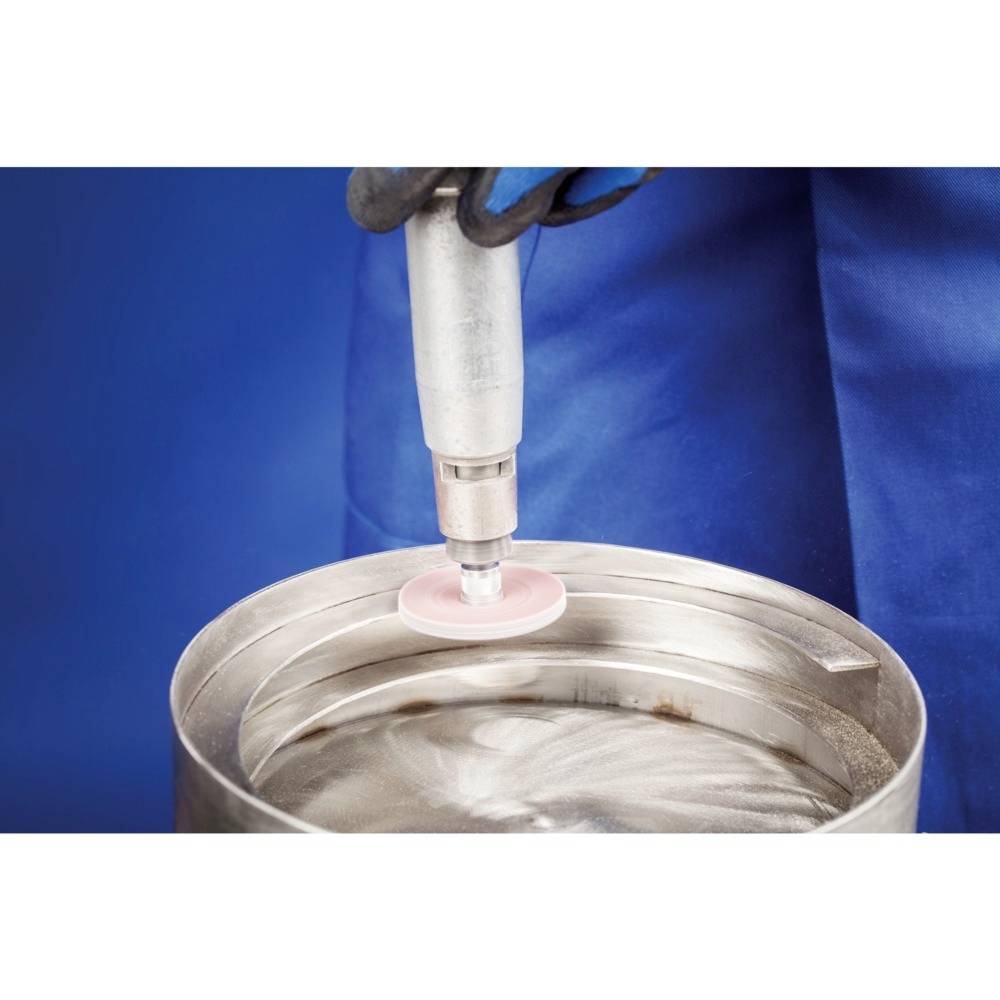 Eine Person schleift die Oberfläche eines Metallfasses mit einem rotierenden Schleifwerkzeug. Sie trägt Handschuhe und einen blauen Schutzanzug.